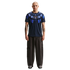 A full-body studio portrait of a bald man with arm tattoos standing against a solid black background. He is wearing a dark navy blue Nike t-shirt featuring a bright blue radial graphic around the collar and a center-chest Uruguay national team crest. His