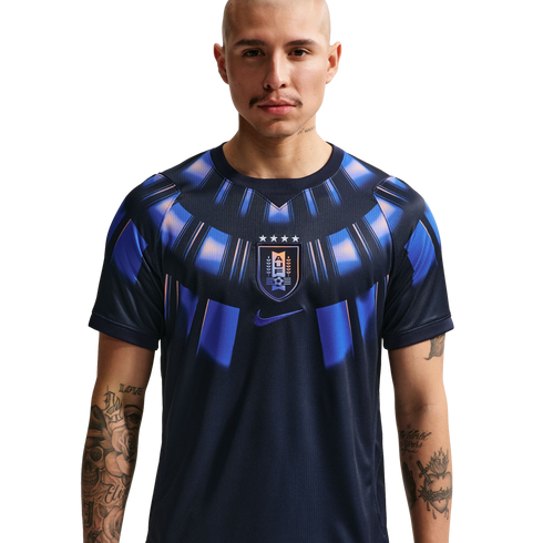 A waist-up studio portrait of a bald man with arm tattoos wearing a dark navy Uruguay national team soccer jersey featuring a blue and orange sunburst pattern around the collar and the AUF crest in the center against a solid black background.