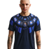 A waist-up studio portrait of a bald man with arm tattoos wearing a dark navy Uruguay national team soccer jersey featuring a blue and orange sunburst pattern around the collar and the AUF crest in the center against a solid black background.