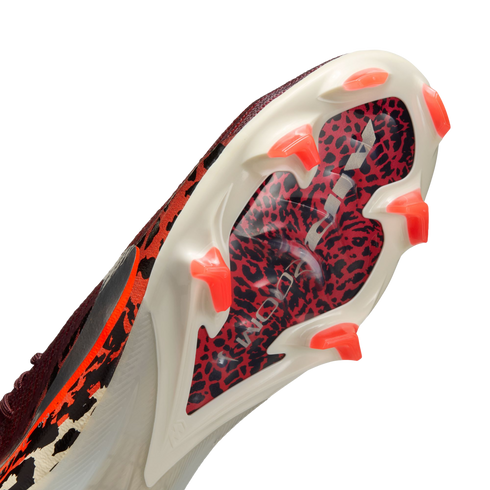 An angled, close-up view of the soleplate of a white soccer cleat, featuring a red and black leopard print pattern under a clear section with silver "AIR ZOOM" branding. The cleat has bright orange, translucent, chevron-shaped studs.