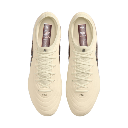 A top-down view of a pair of off-white Nike soccer cleats against a solid black background. The cleats feature cream-colored uppers and laces, with dark reddish-brown insoles containing white text and logos.