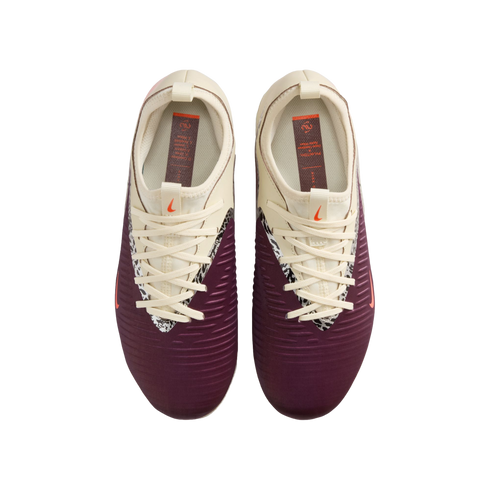 A top-down view of a pair of Nike football boots against a black background. The shoes feature a maroon lower section with horizontal ridges and a cream upper section. A black-and-white patterned trim lines the lace area, and small orange Nike swooshes ar