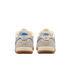 A rear view of a pair of Nike sneakers featuring cream and light gray suede uppers, script Nike logos on distressed blue heel tabs, and gum rubber outsoles against a solid black background.