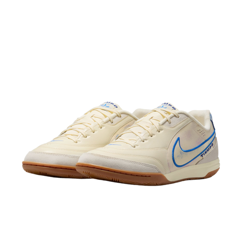 A pair of cream-colored Nike Tiempo indoor soccer shoes with blue accents and gum rubber soles, set against a black background.