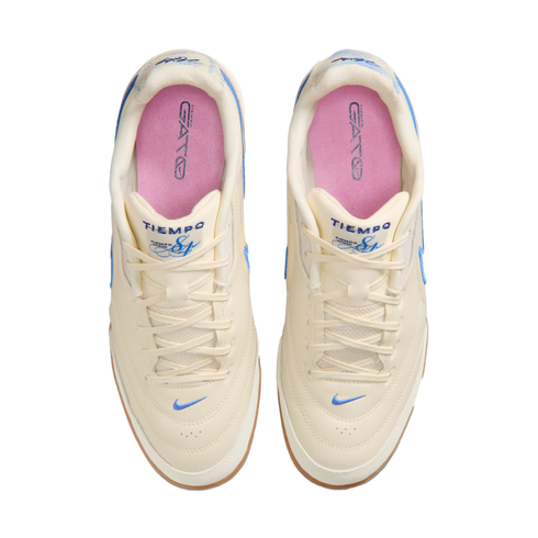 Top-down view of a pair of off-white Nike Tiempo Gato sneakers with light blue accents, pink insoles, and gum rubber outsoles, set against a solid black background.