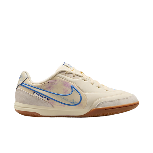 A profile view of a cream-colored Nike Tiempo indoor soccer shoe against a solid black background. The shoe features a blue-outlined Nike swoosh with a faded purple and blue interior, light grey suede overlays on the toe and heel, and a brown gum rubber o