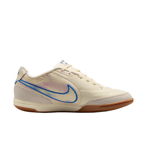 A side profile of a cream-colored Nike indoor soccer shoe against a black background, featuring a blue swoosh with purple accents and a brown gum sole.