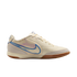 A side profile of a cream-colored Nike indoor soccer shoe against a black background, featuring a blue swoosh with purple accents and a brown gum sole.