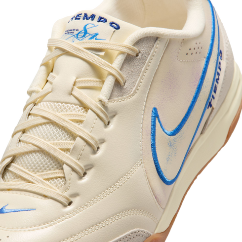 A close-up, angled shot of a cream-colored Nike Tiempo sneaker with blue embroidered accents, matching laces, and subtle iridescent details on the side.