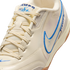 A close-up, angled shot of a cream-colored Nike Tiempo sneaker with blue embroidered accents, matching laces, and subtle iridescent details on the side.