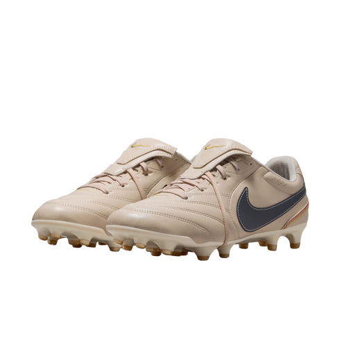 A pair of cream-colored Nike soccer cleats with a dark grey swoosh and gold accents, shown against a solid black background.