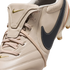 A close-up of a beige Nike soccer cleat featuring a fold-over tongue with a gold logo and a large navy blue side Swoosh with white stitching.