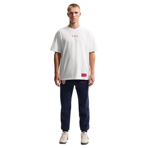 A man stands against a black background wearing a white Nike U.S.A. t-shirt, navy blue USA sweatpants, and white sneakers.