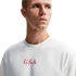 A man in a white Nike crewneck t-shirt with red "U.S.A. NIKE" text on the chest, looking off to the side against a solid black background.