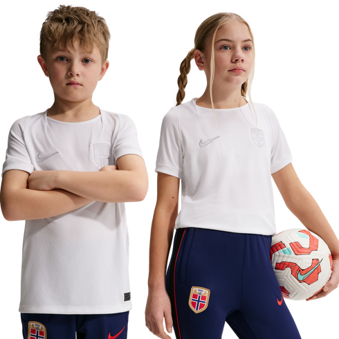 Two children in white Nike soccer jerseys and blue pants with Norway national team crests. The girl holds a soccer ball against a black background.