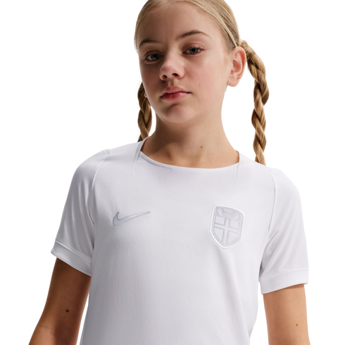 Young girl with blonde pigtail braids wearing a white Nike athletic shirt with a shield crest, posed against a solid black background.