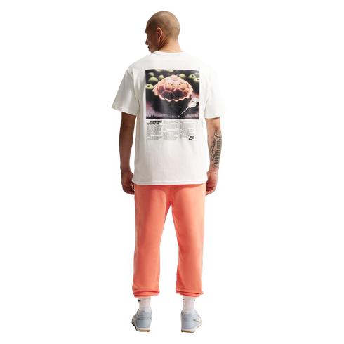 A back view of a man standing against a black background, wearing a white t-shirt with a large graphic of an apple pie and the text "AS AMERICAN AS APPLE PIE." He is also wearing coral-colored sweatpants, white socks, and light blue sneakers. He has a bal