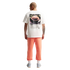 A back view of a man standing against a black background, wearing a white t-shirt with a large graphic of an apple pie and the text "AS AMERICAN AS APPLE PIE." He is also wearing coral-colored sweatpants, white socks, and light blue sneakers. He has a bal