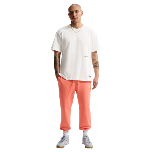 Full-length photo of a bald man with arm tattoos standing against a black background, wearing a white T-shirt, salmon-colored sweatpants, and light blue sneakers.