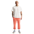 Full-length photo of a bald man with arm tattoos standing against a black background, wearing a white T-shirt, salmon-colored sweatpants, and light blue sneakers.