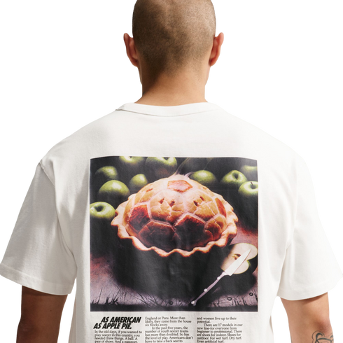 A back view of a person with a shaved head wearing a white t-shirt against a black background. The shirt features a large graphic on the back resembling a vintage magazine advertisement. The main image shows a baked pie with a soccer-ball-patterned crust