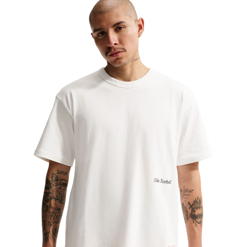 A man with a shaved head and tattooed arms wears a white Nike Football t-shirt against a black background.