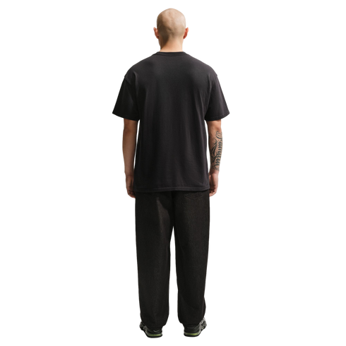 A full-length rear view of a bald man wearing a plain black t-shirt and loose-fitting black pants against a solid black background. He has a large tattoo on his right forearm.