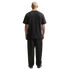 A full-length rear view of a bald man wearing a plain black t-shirt and loose-fitting black pants against a solid black background. He has a large tattoo on his right forearm.