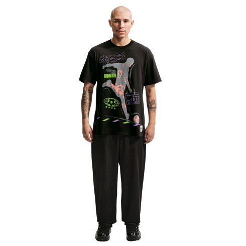 A full-body portrait of a bald man with a mustache and arm tattoos, standing against a solid black background. He is wearing a black graphic t-shirt with a soccer player illustration, loose black trousers, and black sneakers with neon green details.