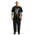 A full-body portrait of a bald man with a mustache and arm tattoos, standing against a solid black background. He is wearing a black graphic t-shirt with a soccer player illustration, loose black trousers, and black sneakers with neon green details.