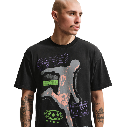 A man with a shaved head and mustache wears a black graphic t-shirt against a solid black background. The shirt features a large grey and peach-colored silhouette of an athlete in motion, surrounded by various purple and green sports-themed logos and text