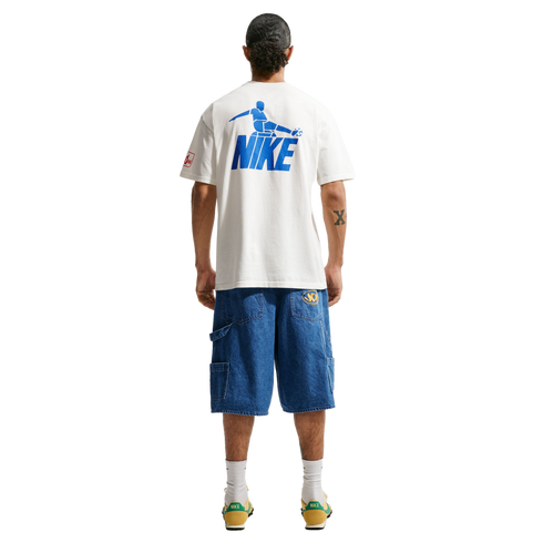 A person stands with their back to the camera against a black background, wearing a white t-shirt featuring a blue Nike soccer graphic on the back, loose-fitting blue denim shorts, white crew socks, and yellow and green sneakers.