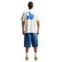 A person stands with their back to the camera against a black background, wearing a white t-shirt featuring a blue Nike soccer graphic on the back, loose-fitting blue denim shorts, white crew socks, and yellow and green sneakers.