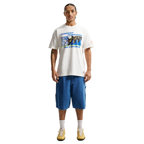 A person stands against a black background wearing a white "Friendship Tournament" Nike graphic t-shirt, knee-length blue denim shorts, white socks, and yellow sneakers.