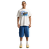 A person stands against a black background wearing a white "Friendship Tournament" Nike graphic t-shirt, knee-length blue denim shorts, white socks, and yellow sneakers.