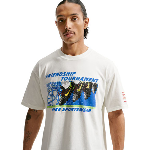 A man with a thin mustache and arm tattoos wears a white t-shirt featuring a colorful graphic of soccer cleats and a ball with the text "Friendship Tournament" and "Nike Sportswear" in blue. He is posed against a solid black background.