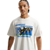 A man with a thin mustache and arm tattoos wears a white t-shirt featuring a colorful graphic of soccer cleats and a ball with the text "Friendship Tournament" and "Nike Sportswear" in blue. He is posed against a solid black background.