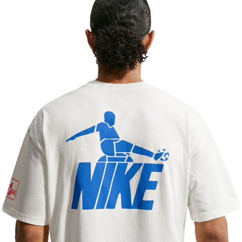 A back view of a person with dark, curly hair tied in a bun, wearing a white t-shirt against a black background. The shirt features a large blue graphic on the back consisting of a stylized soccer player kicking a ball, positioned above the word "NIKE" in