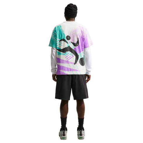 A back view of a person standing against a solid black background, wearing a black beanie, a colorful graphic t-shirt layered over a white long-sleeved shirt, black shorts, and grey sneakers.