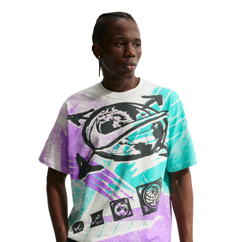 A young Black man with braided hair poses against a solid black background, wearing a white Nike t-shirt with a large, vibrant graphic. The shirt's central design features a black globe with a Nike swoosh wrapped around it, surrounded by bold, textured sp