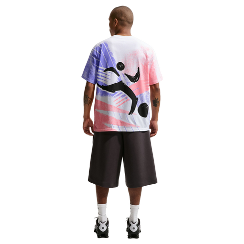 A rear view of a man standing against a black background. He is wearing a white oversized t-shirt with a large pink, purple, and black abstract graphic on the back, dark gray knee-length shorts, white crew socks, and black and white athletic shoes.