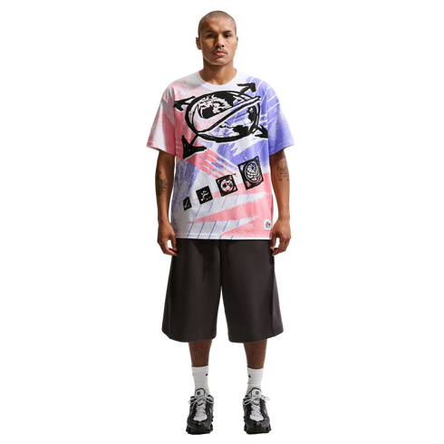 A full-length photo of a man standing against a black background, wearing an oversized graphic t-shirt with pink and purple spray-paint designs, wide-leg charcoal grey shorts, white crew socks, and grey athletic sneakers. He has short hair, a mustache, an