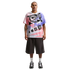 A full-length photo of a man standing against a black background, wearing an oversized graphic t-shirt with pink and purple spray-paint designs, wide-leg charcoal grey shorts, white crew socks, and grey athletic sneakers. He has short hair, a mustache, an