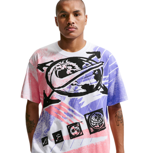 A man with short hair and a mustache stands against a solid black background, wearing a white t-shirt with a large, colorful Nike graphic. The graphic includes a central globe design with a black swoosh, surrounded by arrows and spray-paint style patterns