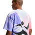 A back view of a person wearing a white t-shirt with a large, abstract graphic. The design features textured purple and pink washes with a dynamic black brushstroke figure in the center.