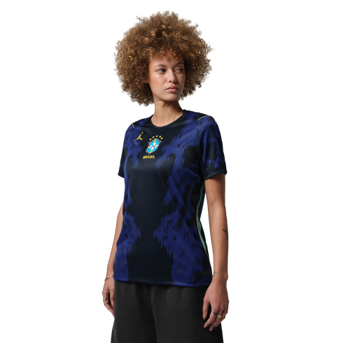 A person with a large curly afro wearing a blue and black patterned Brazil national team jersey, looking to the side against a black background.