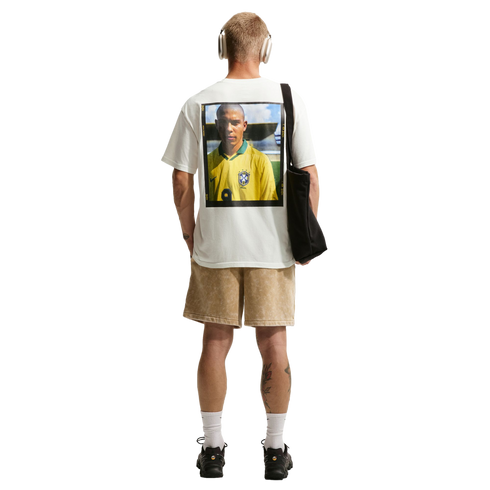A rear view of a man with short blonde hair standing against a solid black background. He wears a white t-shirt with a large graphic print of a soccer player in a yellow Brazil jersey on the back, paired with tan shorts, white crew socks, and black sneake