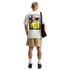A rear view of a man with short blonde hair standing against a solid black background. He wears a white t-shirt with a large graphic print of a soccer player in a yellow Brazil jersey on the back, paired with tan shorts, white crew socks, and black sneake