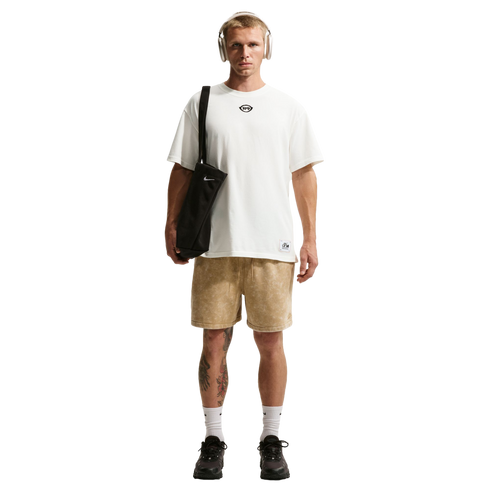 A man with short blonde hair and tan headphones stands against a black background, wearing an oversized white t-shirt and light brown patterned shorts. He carries a black Nike tote bag over his shoulder and wears white socks with black sneakers. Tattoos a