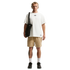 A man with short blonde hair and tan headphones stands against a black background, wearing an oversized white t-shirt and light brown patterned shorts. He carries a black Nike tote bag over his shoulder and wears white socks with black sneakers. Tattoos a